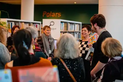 Dans les rayons de la Bibliothèque, une autrice parle en souriant à une assemblée de personnes debout.