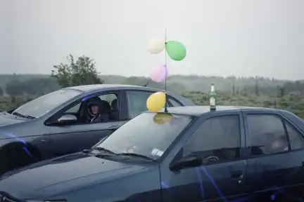 La photographie représente deux véhicules stationnés dans la nature. Le temps est brumeux. La première voiture est décorée avec des ballons de baudruche colorés. Une bouteille est posée sur le toit. Un homme avec une capuche est assis à la place conducteur de la seconde voiture.