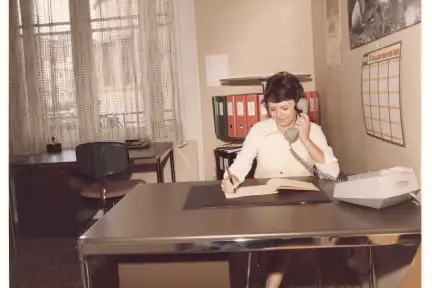 Une jeune femme en tenue de travail de la poudrerie pose devant une toile peinte. Elle est vêtue d'un béret, et d'une combinaison dont les jambes s'arrêtent aux genoux.