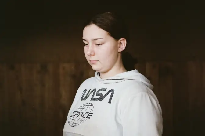 Liya Une jeune femme photographiée en buste, orientée de trois quart vers la gauche, porte son regard vers le bas de l'image. Elle se détache du fond par son visage et son sweat de couleur blanc et gris. son expression est douce. C'est une photographie couleur