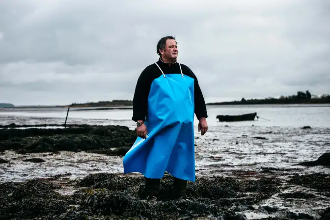 : Au premier plan de cette photographie qui est prise depuis la plage, se trouve un homme, tourné de 3/4 vers le photographe, qui regarde l'horizon vers la droite de la photo. Il porte un tablier de travail bleu, qui capte le regard. Il se tient debout, face au vent le pied dans le goëmon. Sa posture donne un sentiment de force. En arrière-plan on voit un bras de mer et la côte. Photographie en couleur