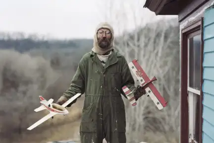Gathered leaves Un homme debout dans la neige, à côté d'une maison, tient un avion en jouet dans chaque main