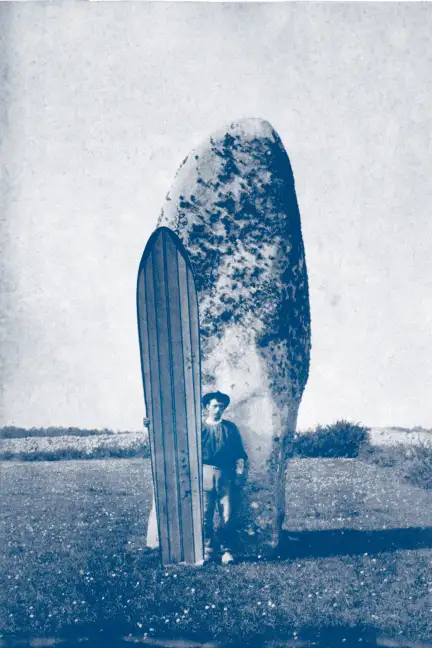 Et si, le surf trouvait ses sources ici … Dans un champ, un homme se tient debout au centre de l'image, devant un menhir d'une hauteur d'environ 5m. En arrière plan on devine un paysage de landes. Il tient dans sa main droit une planche de surf, longue de deux fois sa taille.