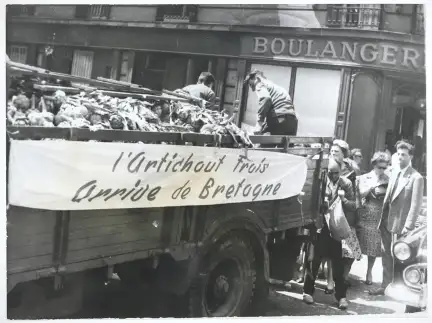 Photographie d'une vente sauvage d’artichauts à Paris. Une camionnette avec une banderole "L'artichaut Frais / Arrive de Bretagne" est au centre de l'image.